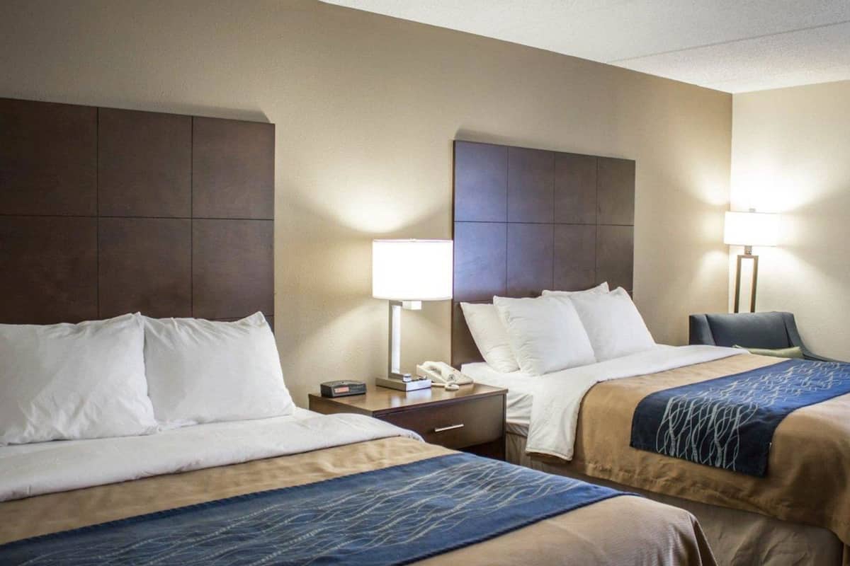 A hotel room with two beds, each with a headboard, bedside tables with lamps, and a chair.