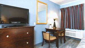 A hotel room with a flat-screen TV, a wooden dresser, a dining table with chairs, a mirror, and a window with striped curtains.