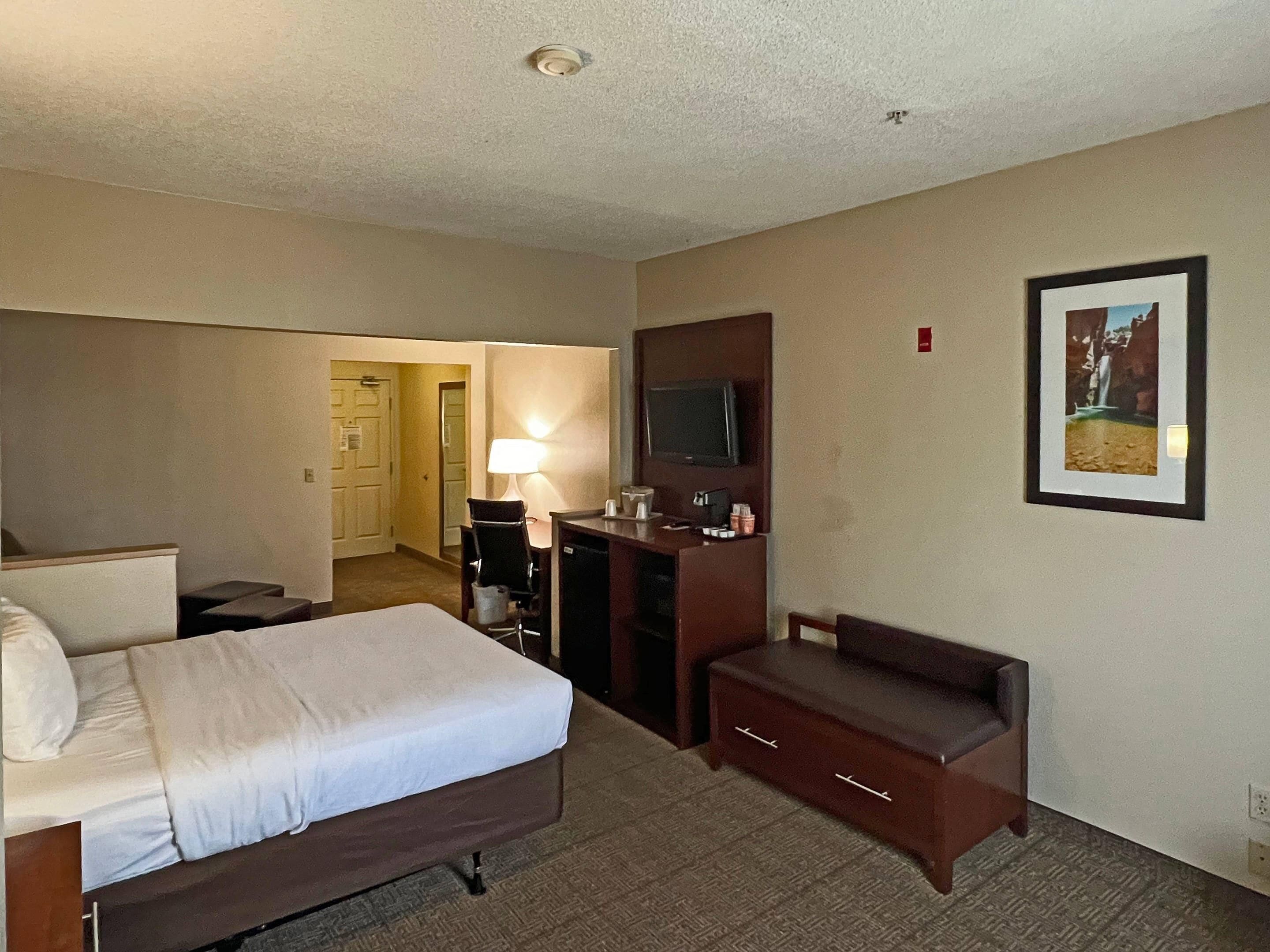 A hotel room with a bed, a desk with a TV, a chair, a lamp, and a framed picture on the wall.