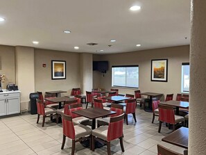 A hotel dining area with multiple tables and chairs, a kitchen area, and large windows providing natural light. - Comfort Suites Peoria Sports Complex (Peoria)