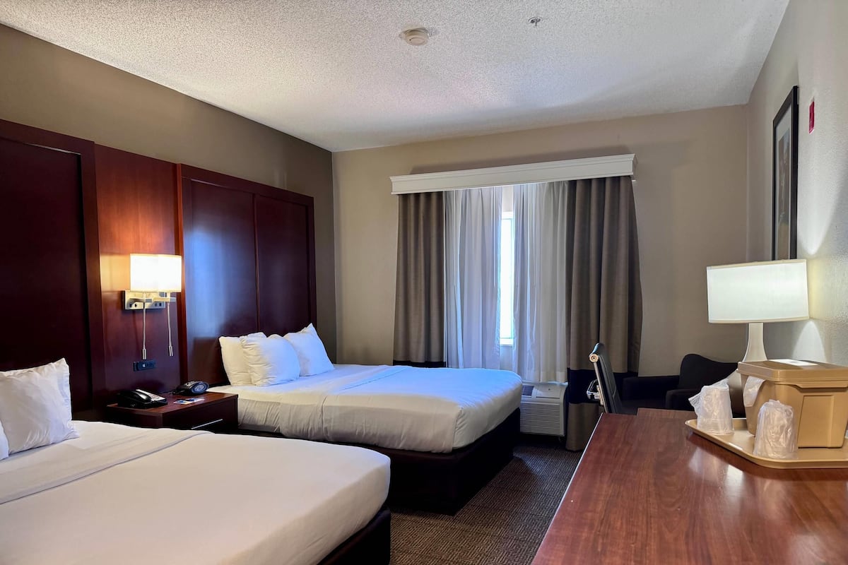 A hotel room with two beds, a desk, a chair, a desk lamp, a telephone, and a wall-mounted TV.