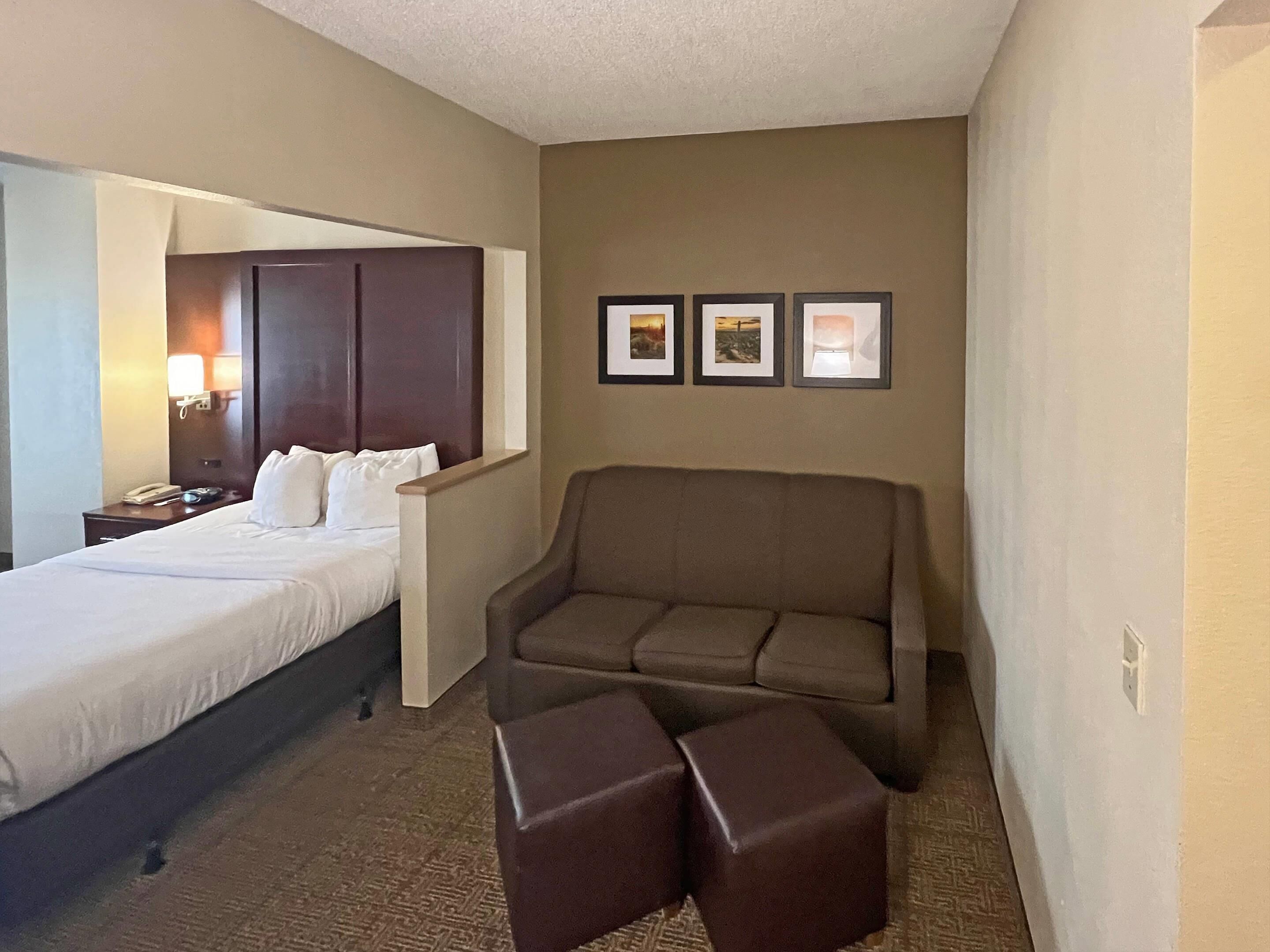 Habitación de hotel con una cama, un sofá marrón, dos otomanas, una mesita de noche con un teléfono y cuadros en la pared.