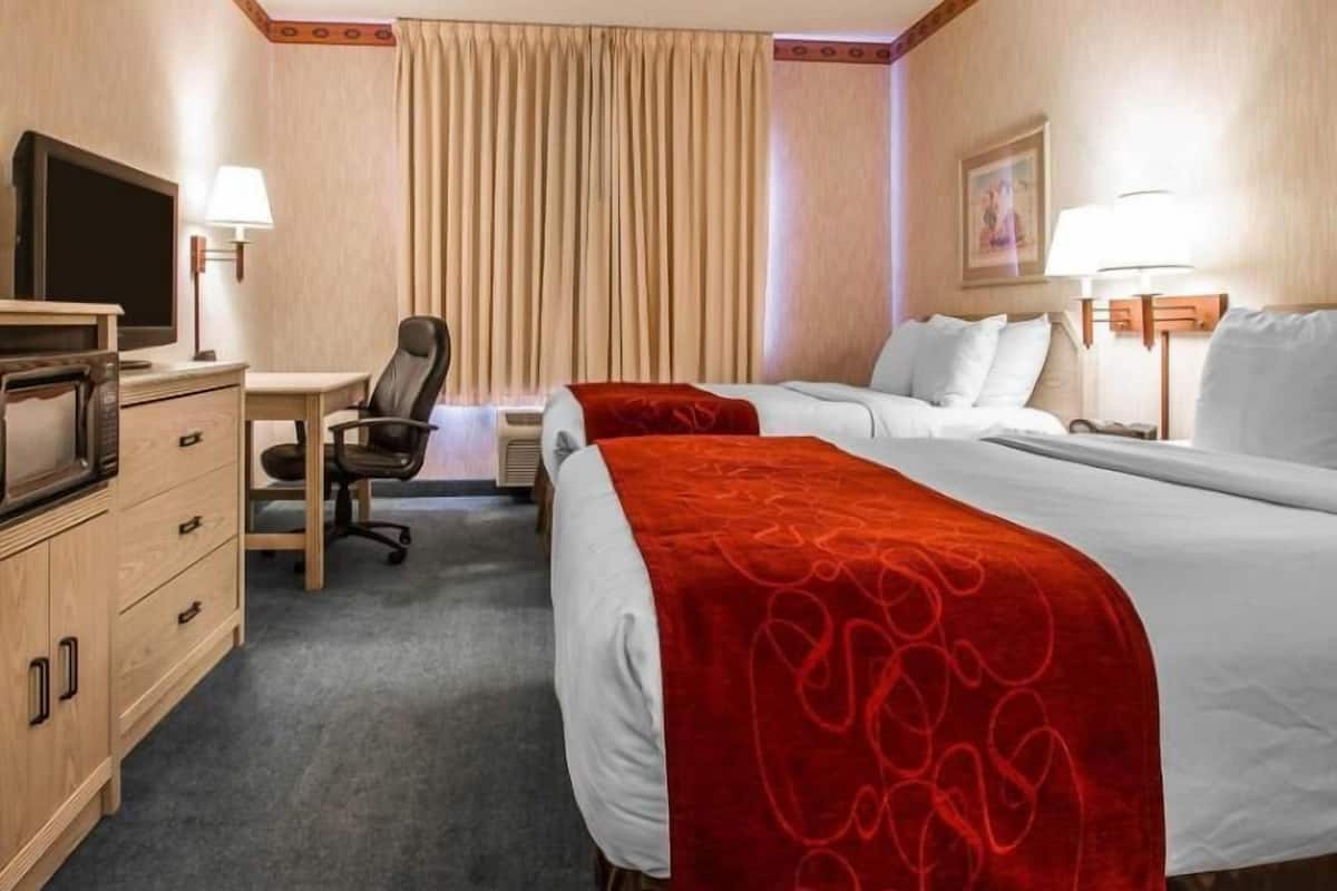 A hotel room with two beds, a desk, a chair, a TV, and a lamp.