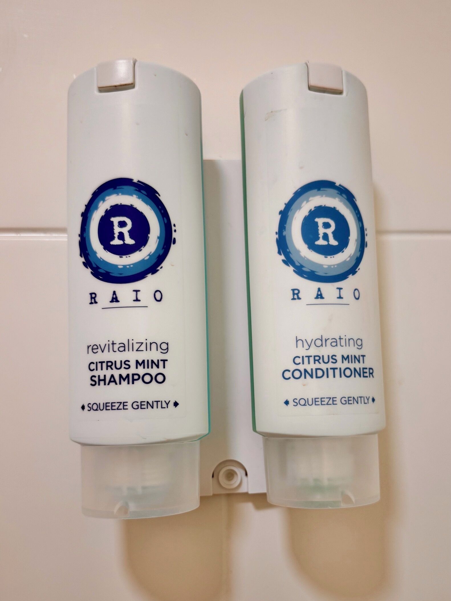 Deux flacons de produits de soins capillaires RAIO, l’un contenant du shampooing et l’autre de l’après-shampooing, avec la consigne de les presser doucement.