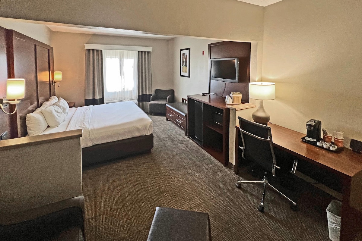 A hotel room with a bed, desk, chair, TV, and a small table with a lamp.