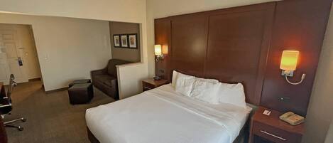 A hotel room with a large bed, a desk, a chair, and a nightstand with a lamp.