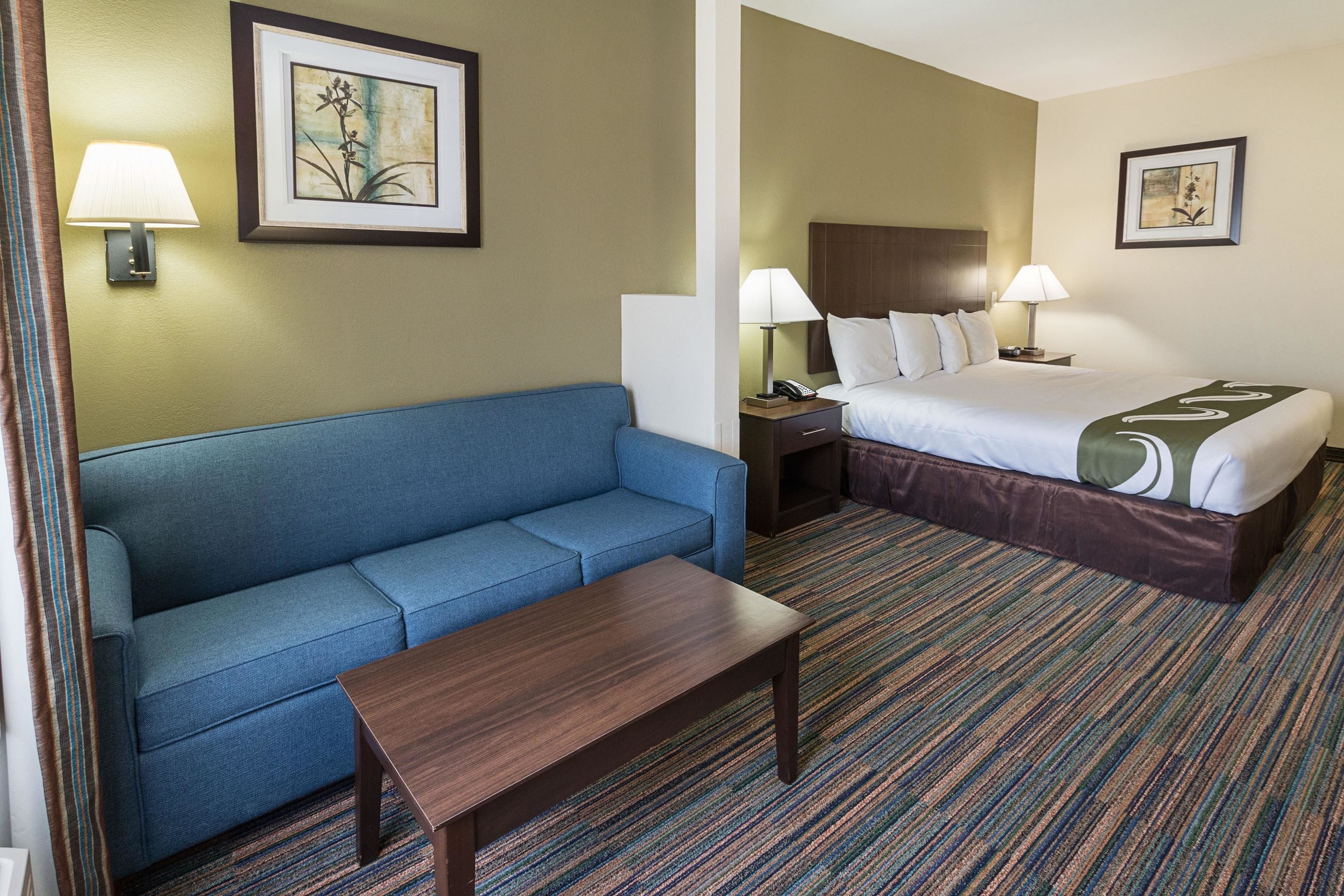 Quarto de hotel com um sofá azul, uma mesa de centro de madeira, uma cama com cabeceira marrom e duas mesas de cabeceira com luminárias.