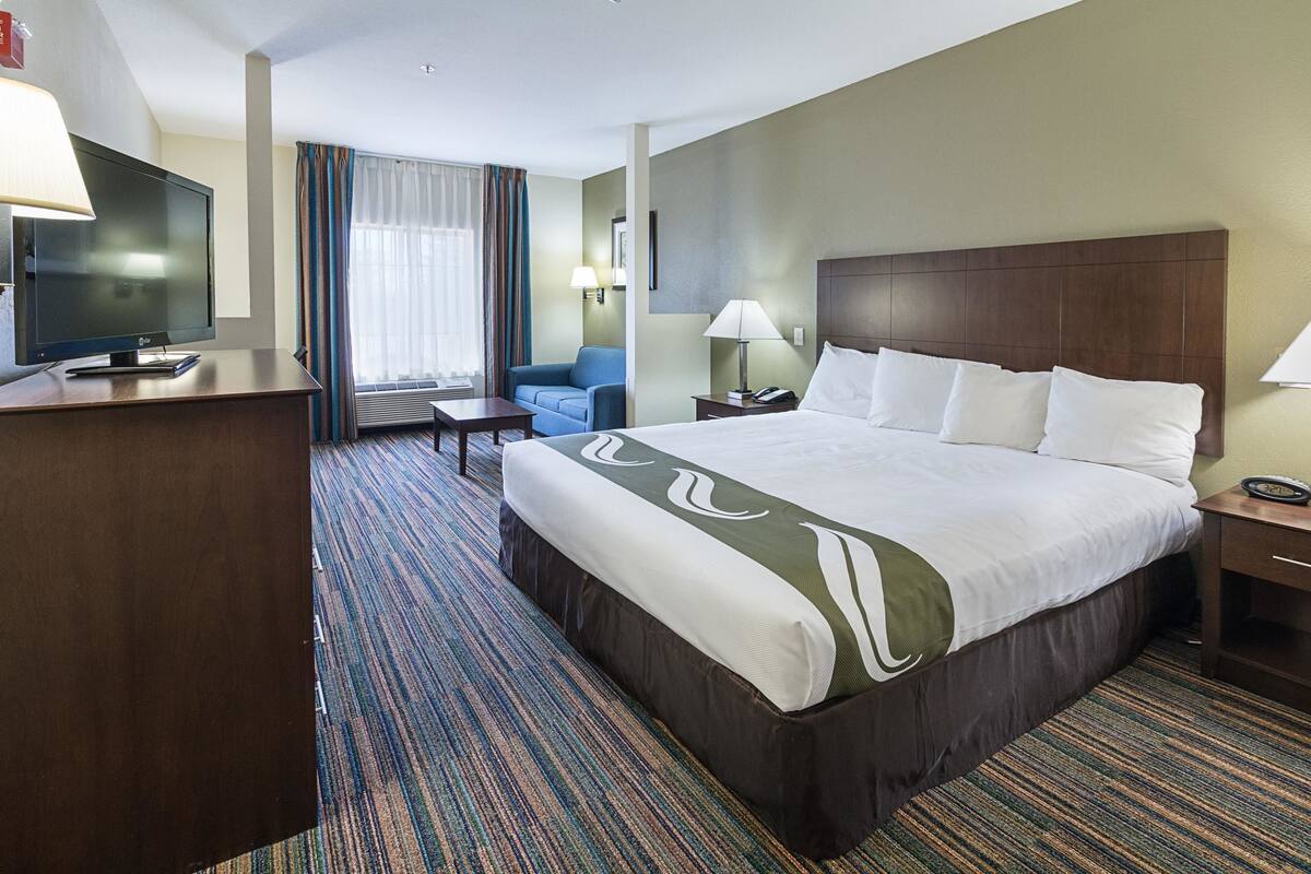 A hotel room with a large bed, a desk with a TV, a chair, a lamp, and a window with curtains.