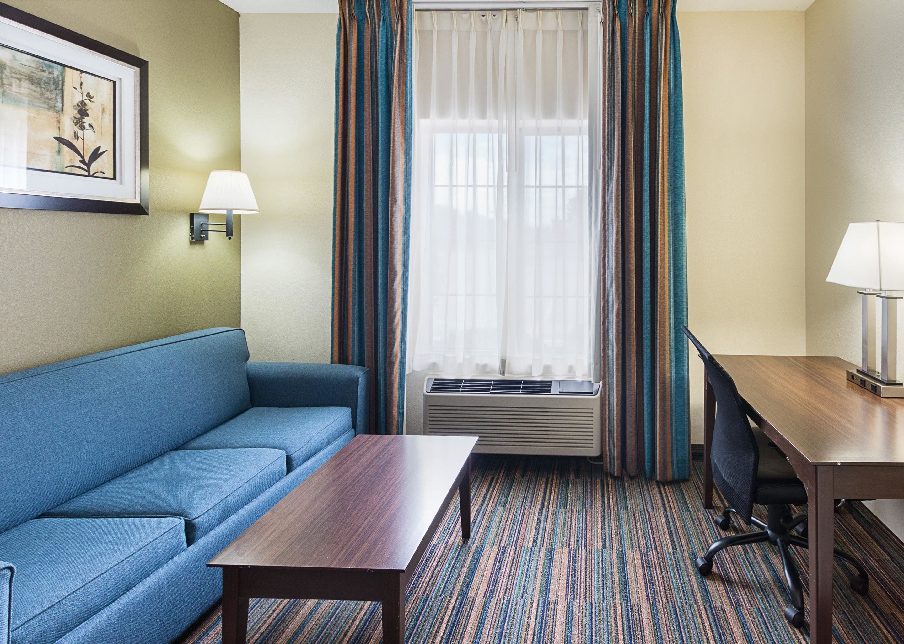 Habitación de hotel con un sofá azul, una mesa de madera, un escritorio con una silla, una ventana con cortinas y una lámpara fijada a la pared.