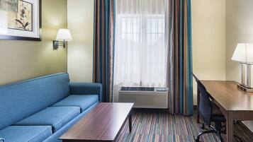 Habitación de hotel con un sofá azul, una mesa de madera, un escritorio con una silla, una ventana con cortinas y una lámpara fijada a la pared.