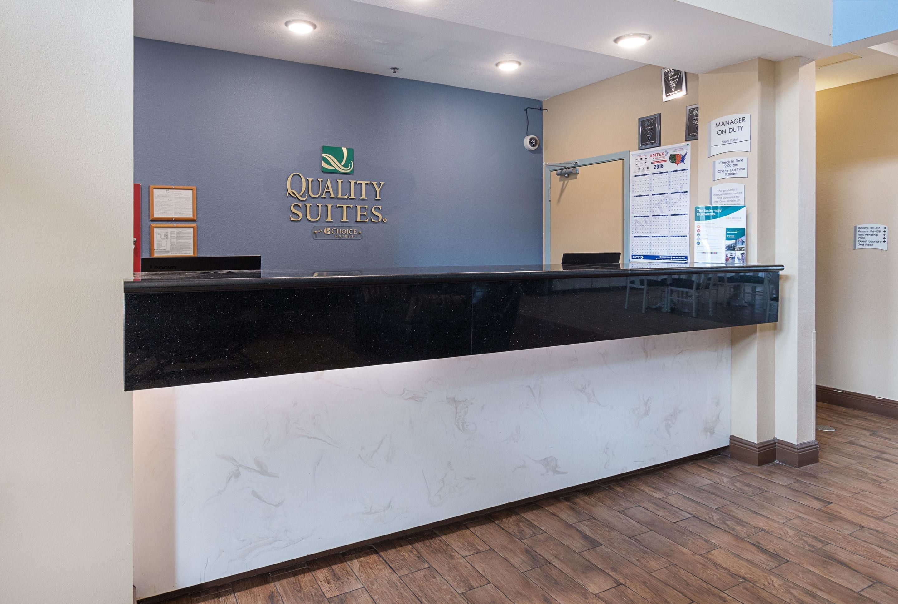 Un mostrador de recepción con diseño de mármol, una pared azul con el logotipo de "Quality Suites" y un cartel que indica "Gerente de Guardia".