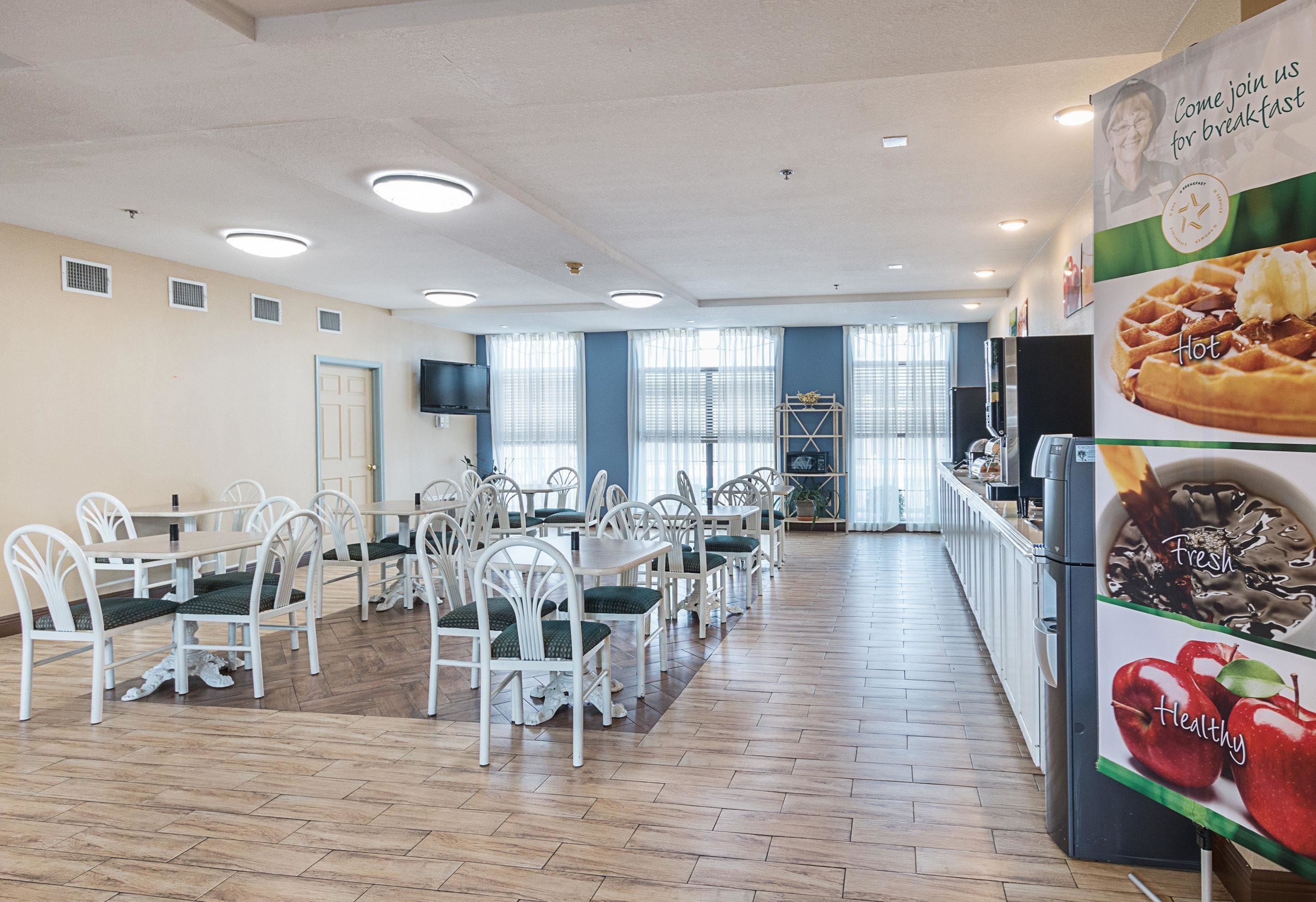 A spacious dining area with white chairs and tables, a large window with blinds, and a wall-mounted television.