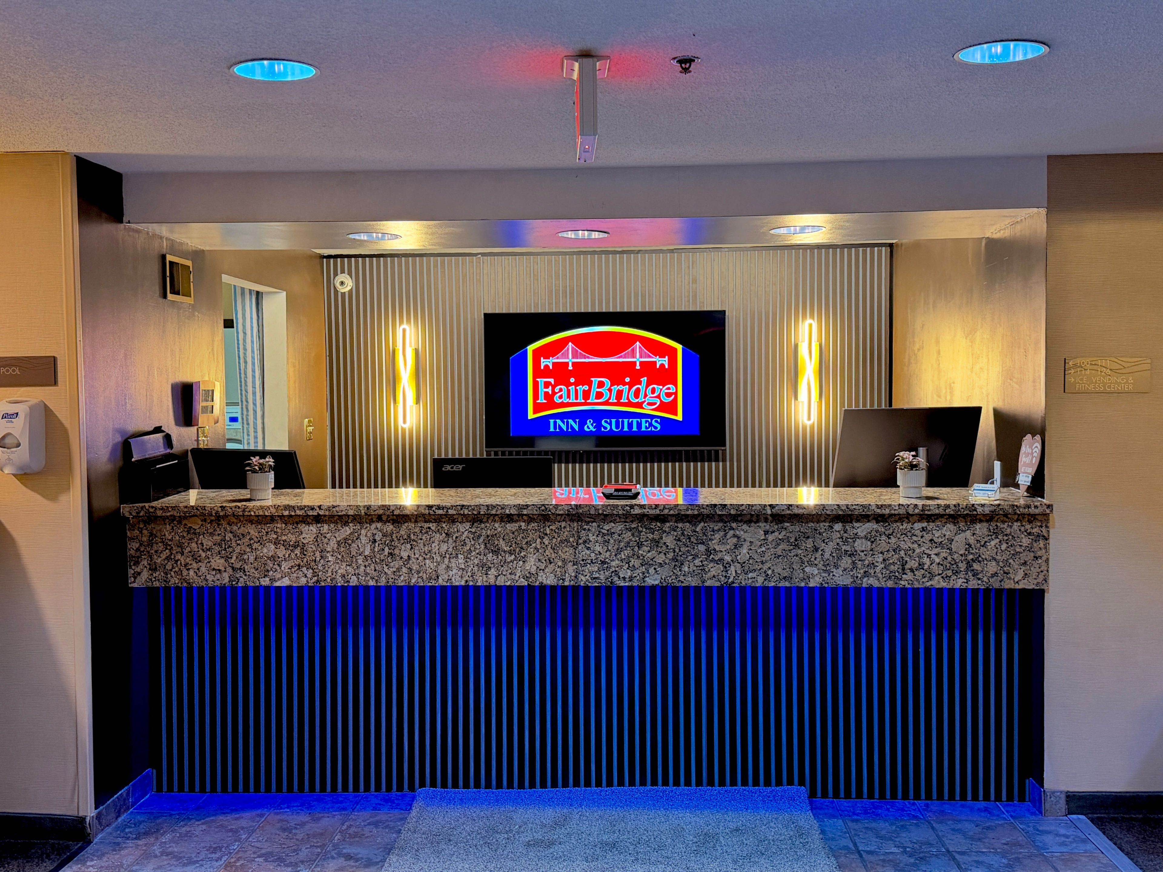 A hotel reception desk with a digital screen displaying the Fairbridge Inn & Suites logo, a reception area with a marble counter, and a blue-lit reception desk.
