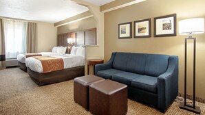 Une chambre d’hôtel comprenant un lit, un canapé, deux poufs, une lampe sur pied et trois tableaux encadrés accrochés au mur.