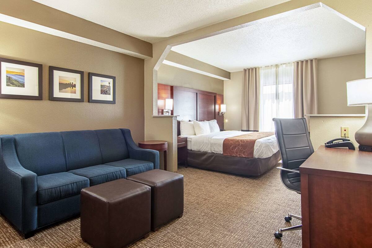 A hotel room with a blue sofa, a bed, a desk, and a chair. There are framed pictures on the wall and a window with curtains.