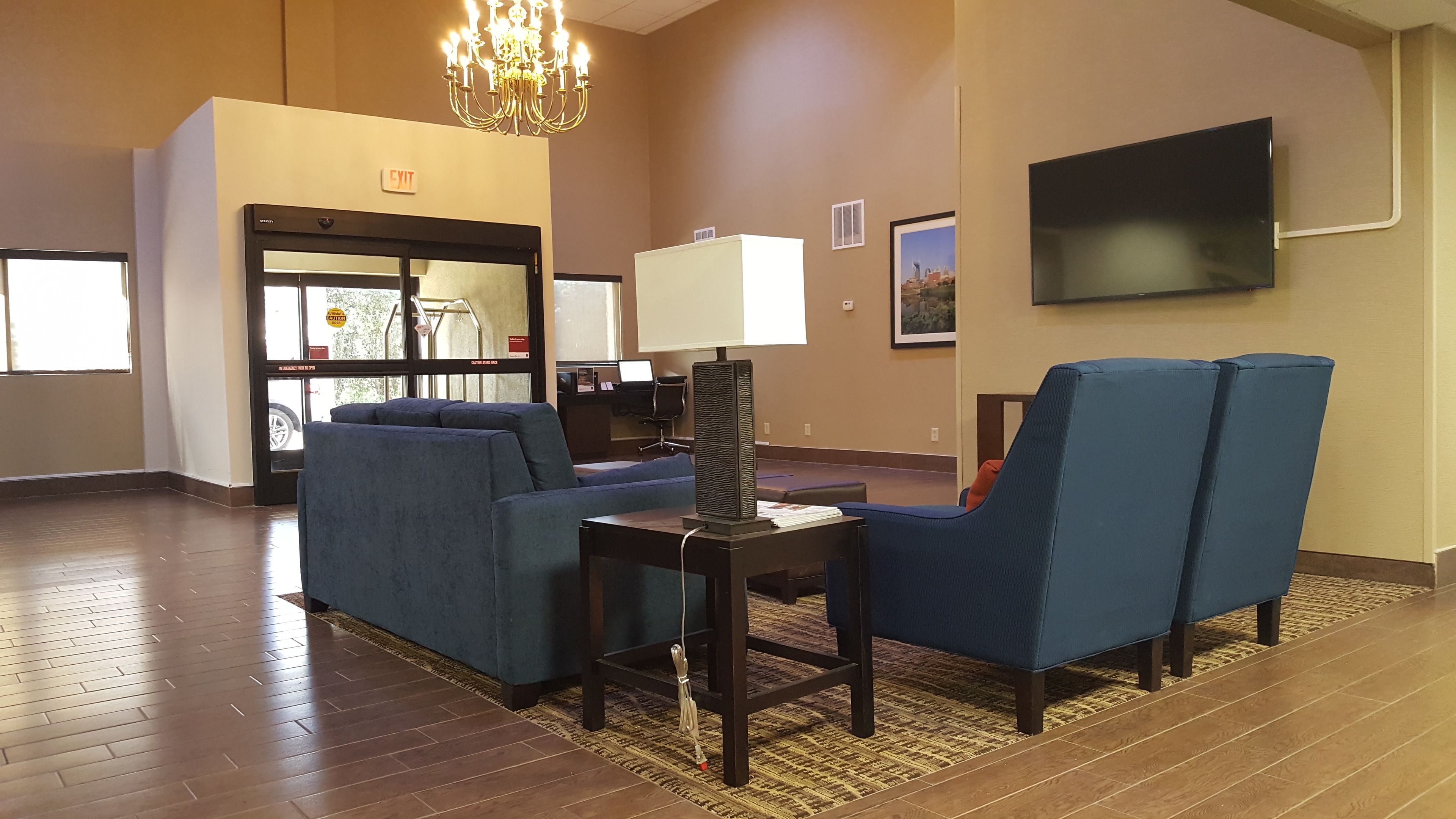 A hotel lobby with a chandelier, a television, blue armchairs, a table with a lamp, and a carpeted floor.