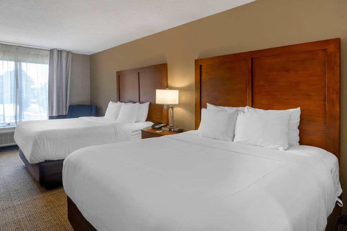 A hotel room with two beds, a wooden headboard, a bedside table with a lamp, and a window with curtains.