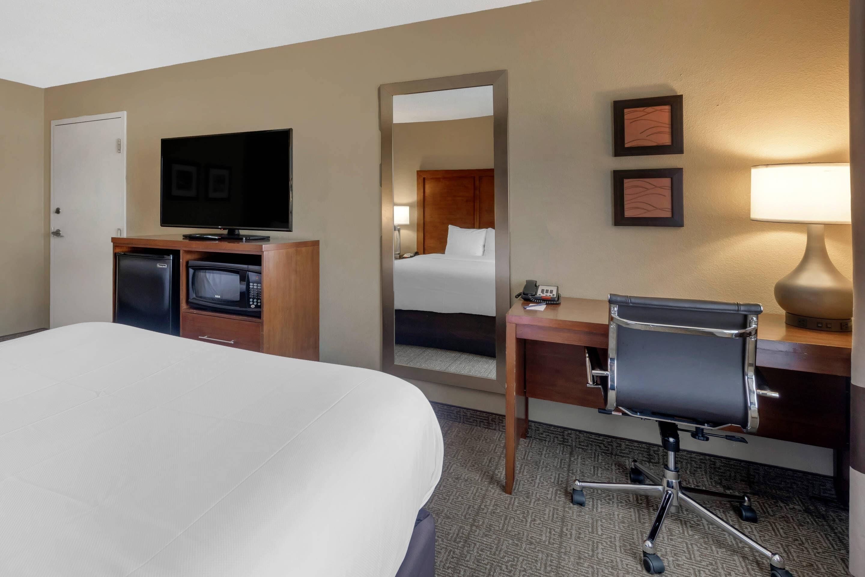 Habitación de hotel con una cama grande, un escritorio con silla, televisión, lámpara de escritorio, microondas y refrigerador.