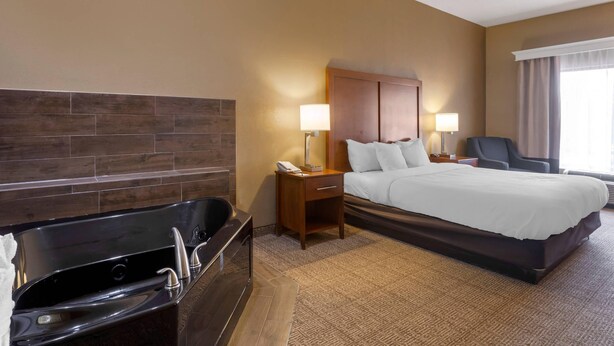 A hotel room with a large bed, a separate bathroom with a bathtub, two bedside lamps, a wooden headboard, and a window with curtains. - Comfort Inn & Suites Peachtree Corners - Norcross (Norcross)