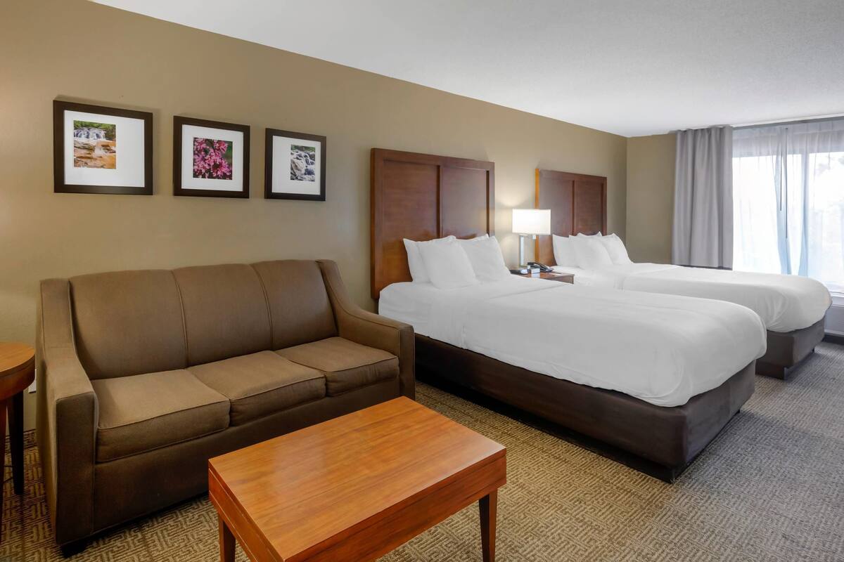 A hotel room with a sofa, two beds, a wooden table, and framed pictures on the wall.