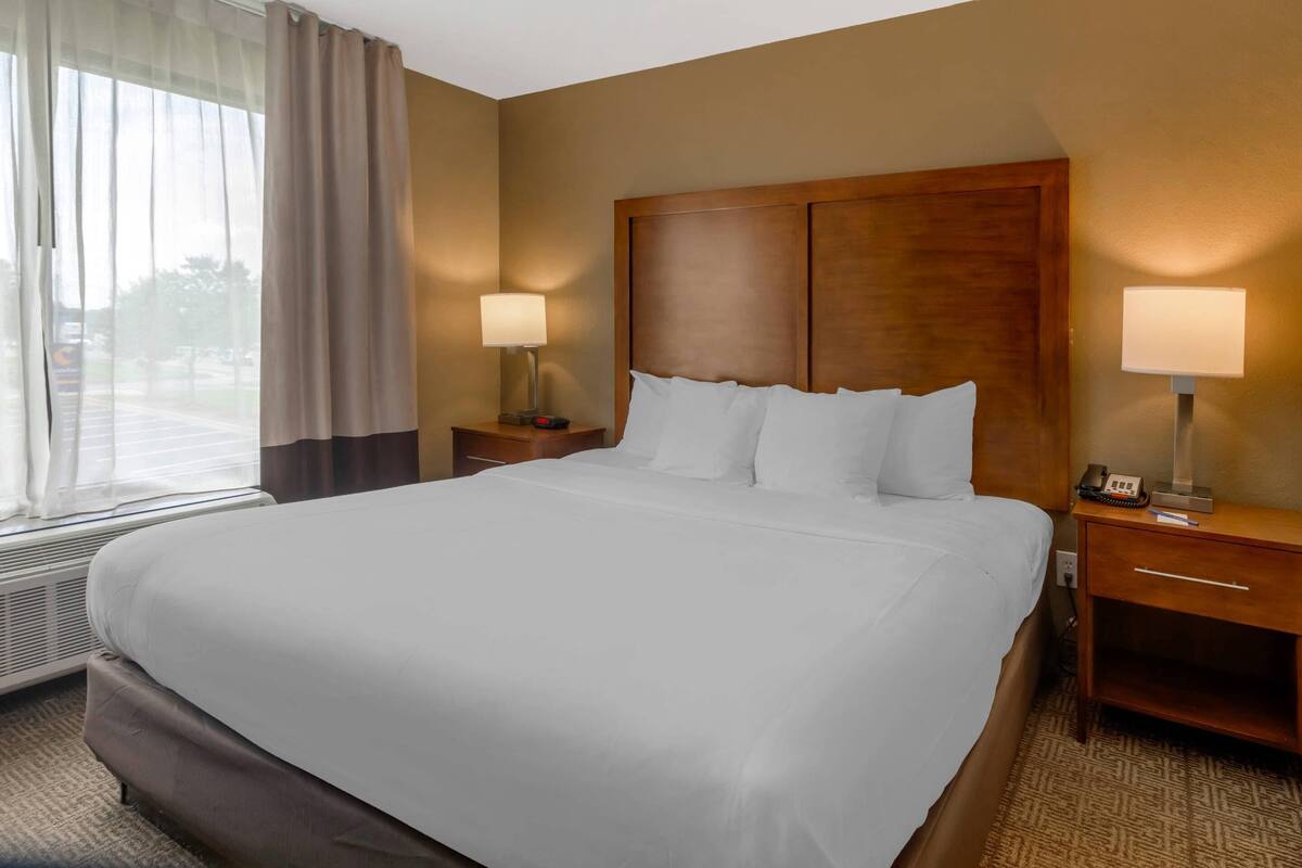 A hotel room with a large bed, two bedside tables with lamps, a window with curtains, and a wall-mounted heater.