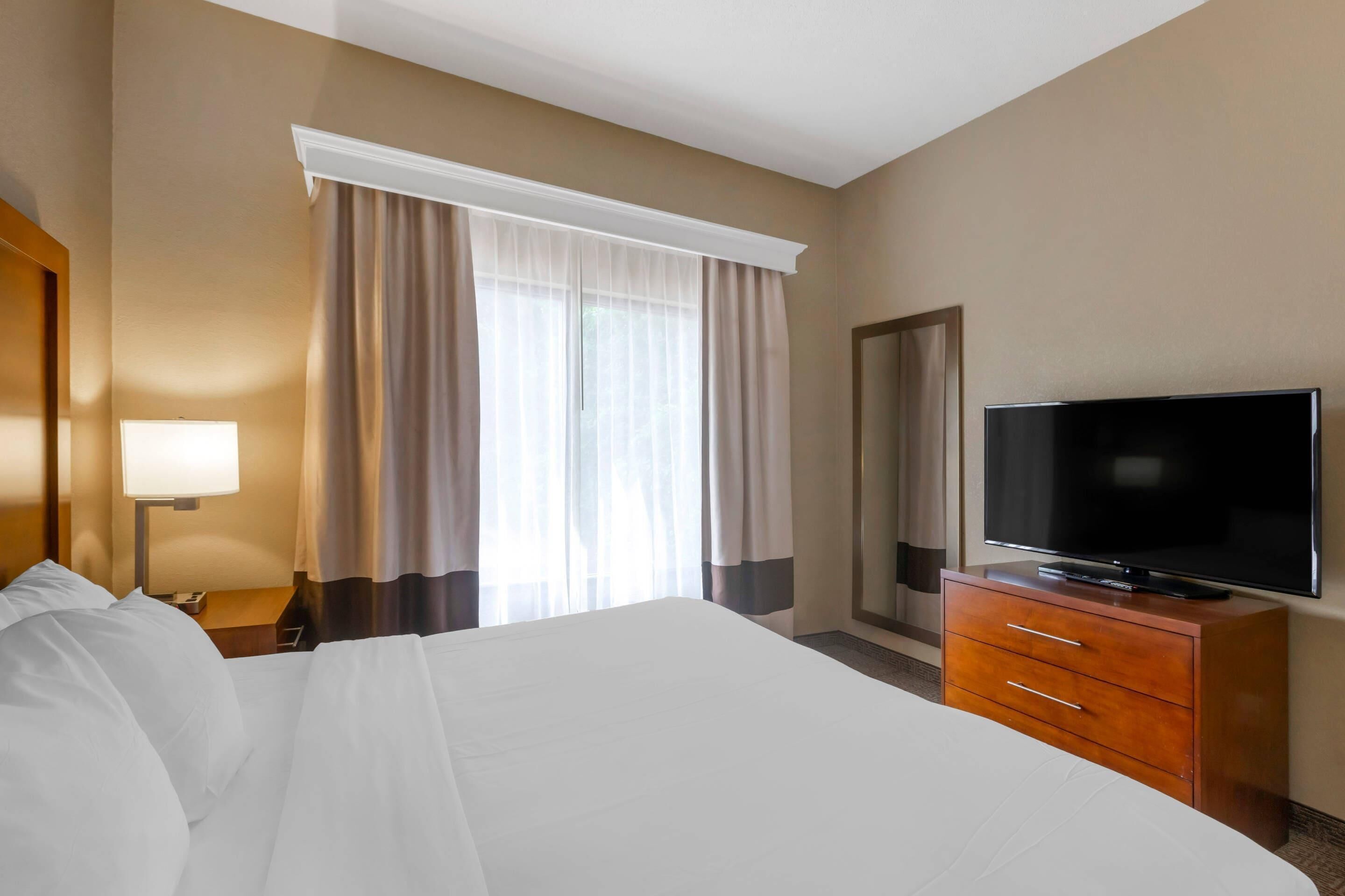 A hotel room with a large bed, a television on a wooden dresser, a nightstand with a lamp, and a window with curtains.