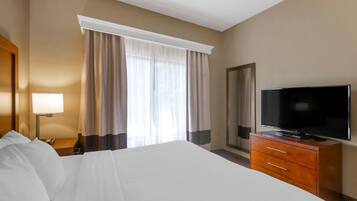 A hotel room with a large bed, a television on a wooden dresser, a nightstand with a lamp, and a window with curtains.