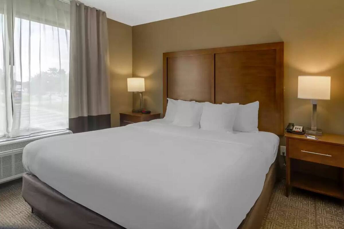 A hotel room with a large bed, two bedside tables with lamps, a window with curtains, and a wall-mounted heater.