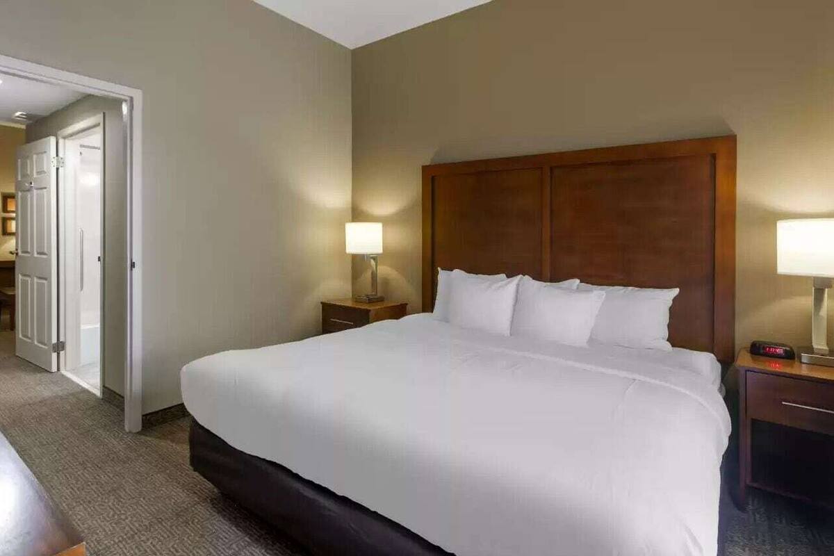 A hotel room with a large bed, a bedside table with a lamp, a nightstand with a clock, and a door leading to another room.