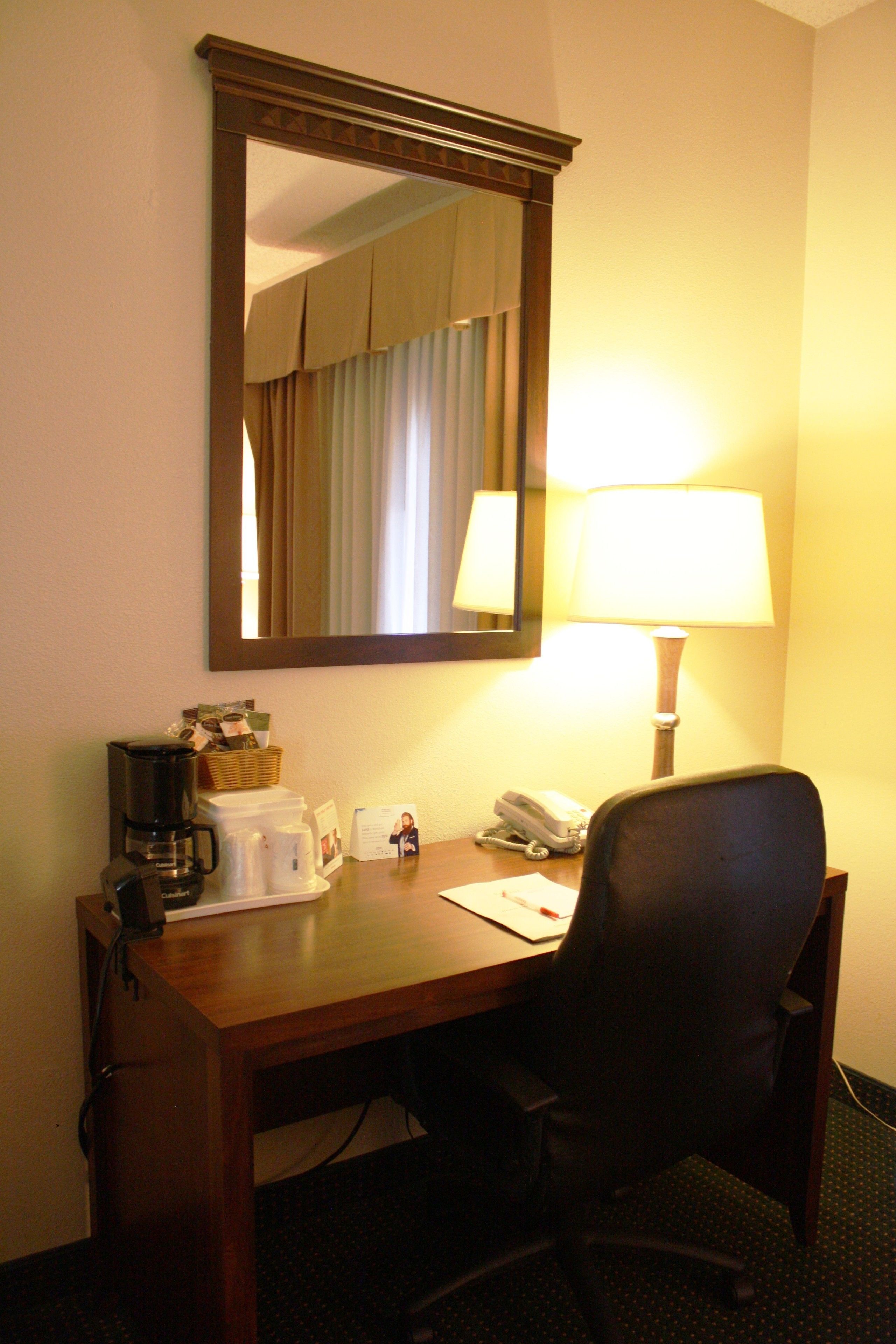A hotel room desk with a lamp, a telephone, a coffee maker, and a mirror.