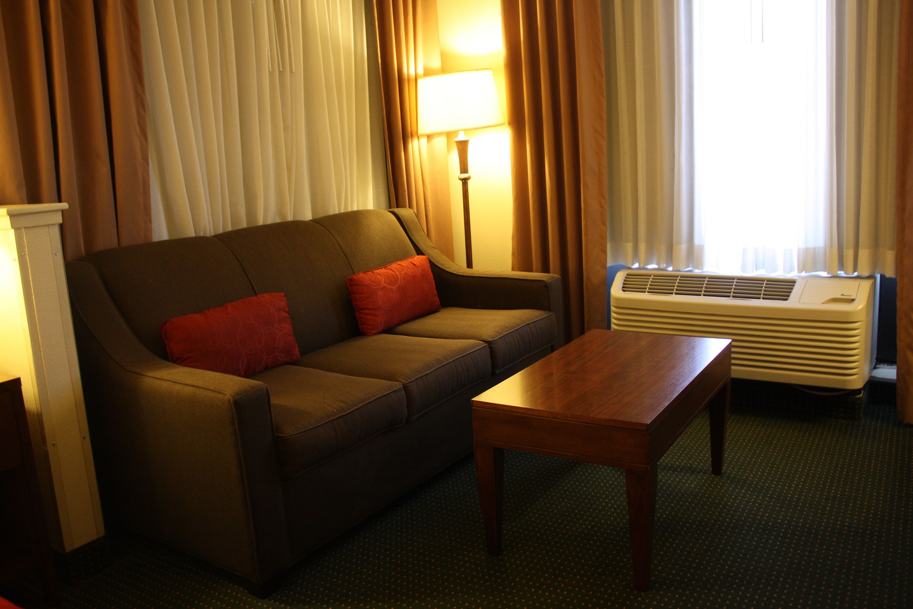 A hotel room with a sofa, a wooden table, a lamp, and an air conditioning unit.
