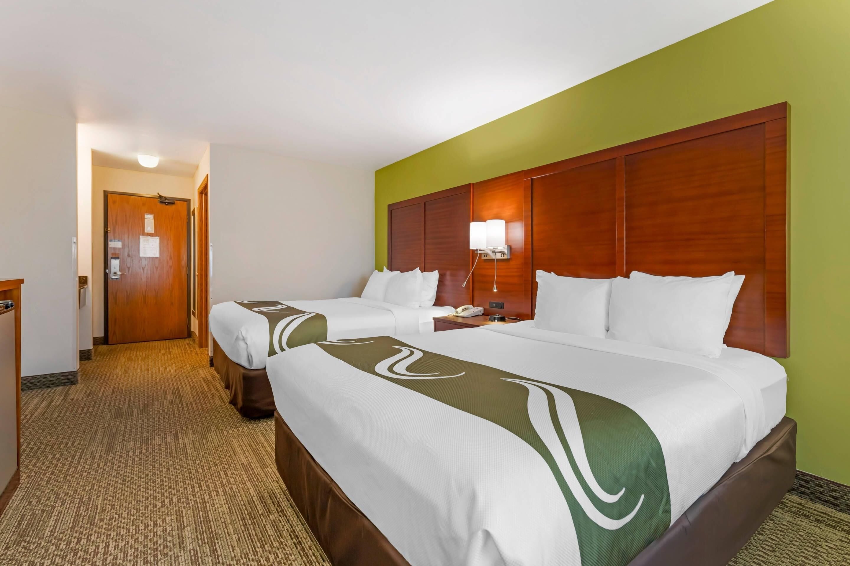 Ein Hotelzimmer mit zwei Betten, einem hölzernen Kopfteil, einem Nachttisch mit Lampe und einem Teppichboden.