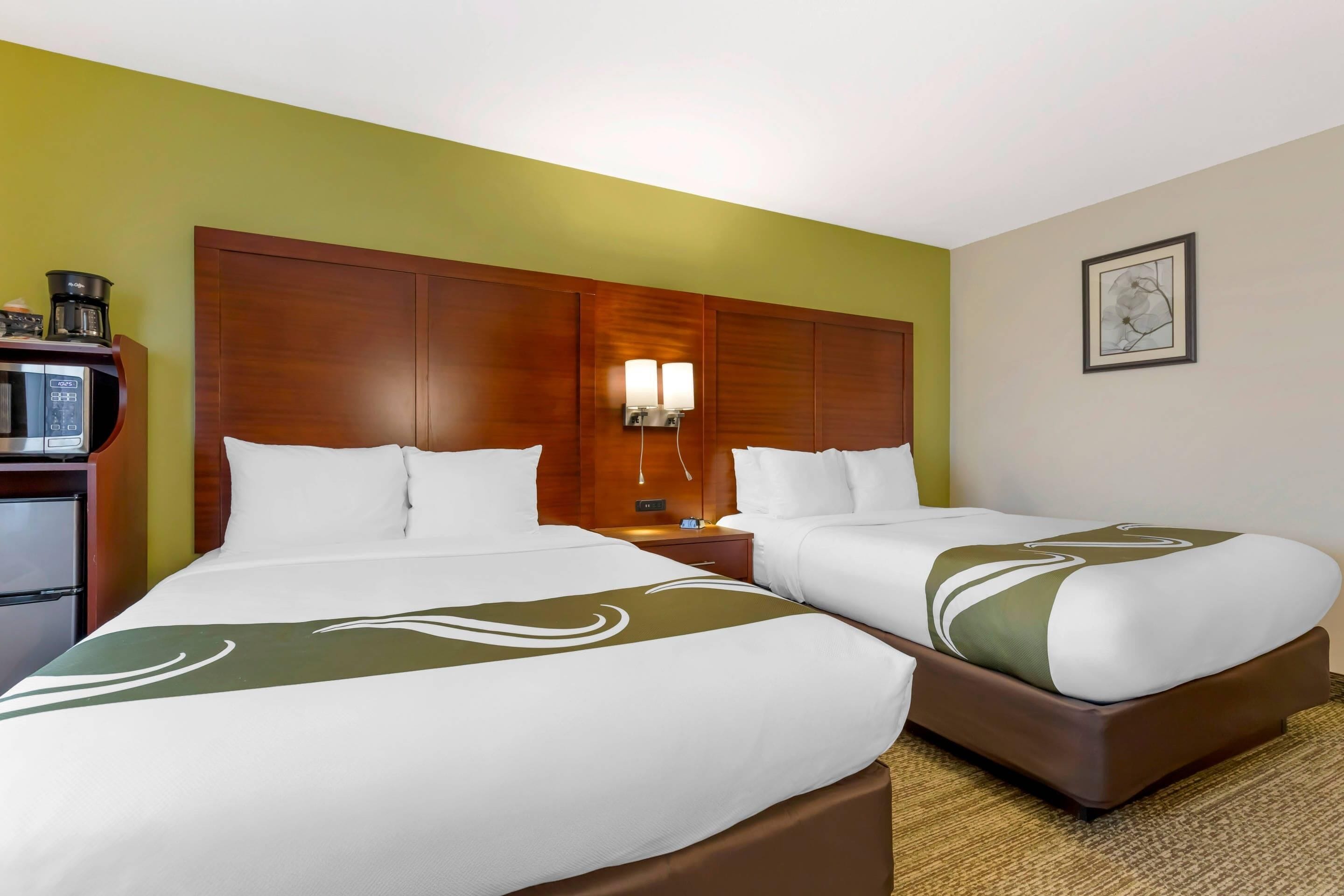 A hotel room with two beds, a wooden headboard, a nightstand with a lamp, a coffee maker, and a framed picture on the wall.