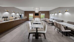 A modern dining area with long tables and chairs, a counter with food and drink options, and a television mounted on the wall.