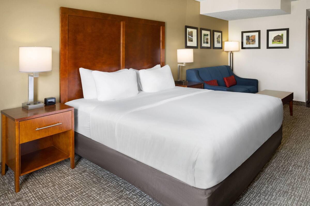 A hotel room with a large bed, a wooden headboard, a nightstand with a lamp, a blue armchair, and a small table.