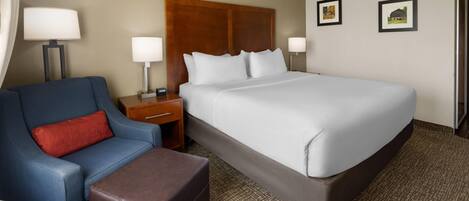 A hotel room with a large bed, a blue armchair, a brown ottoman, two bedside tables with lamps, and two framed pictures on the wall.