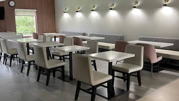 Interior de un restaurante moderno con mesas y sillas blancas, iluminación empotrada en el techo y un amplio ventanal que deja entrar luz natural.