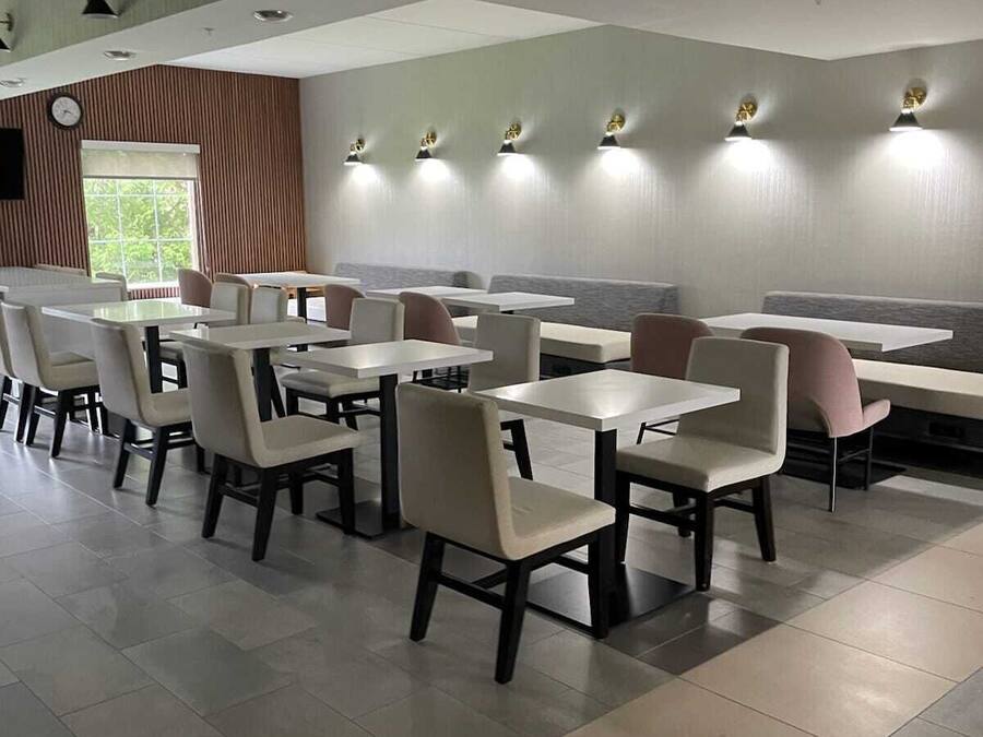 Interior de un restaurante moderno con mesas y sillas blancas, iluminación empotrada en el techo y un amplio ventanal que deja entrar luz natural.