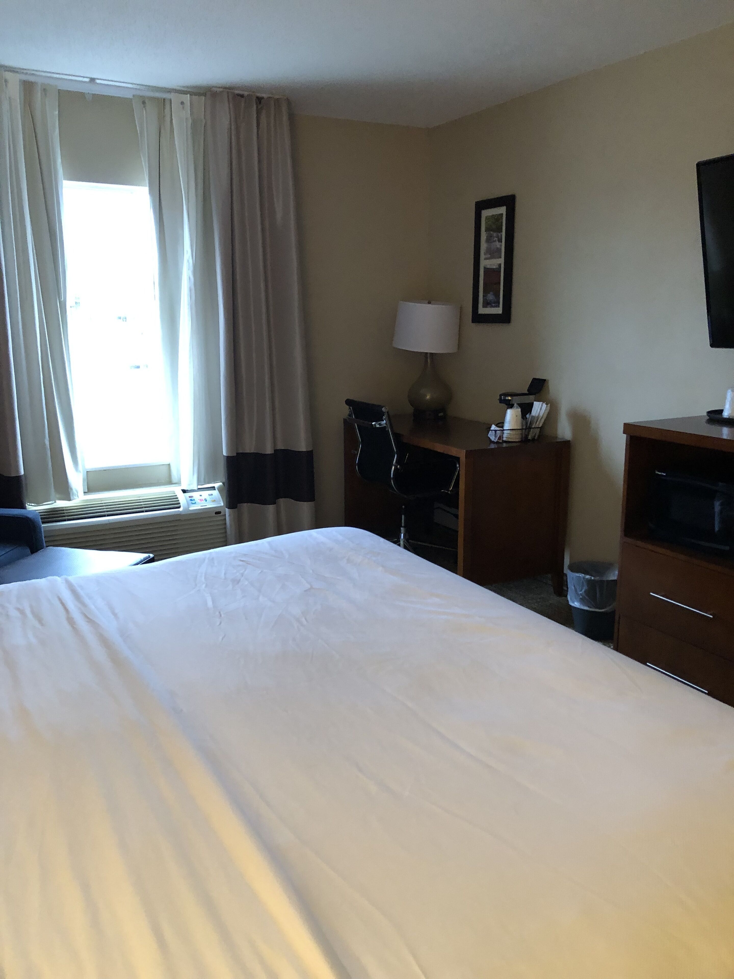 Une chambre d’hôtel avec un grand lit, un bureau avec une lampe, une chaise, une télévision et une fenêtre avec des rideaux.