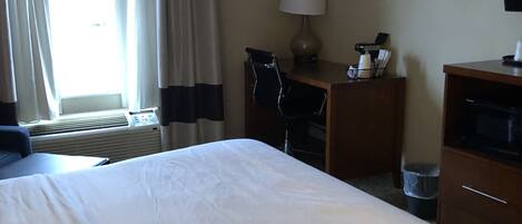 Habitación de hotel con una cama grande, un escritorio con lámpara, una silla, un televisor y un ventanal con cortinas.