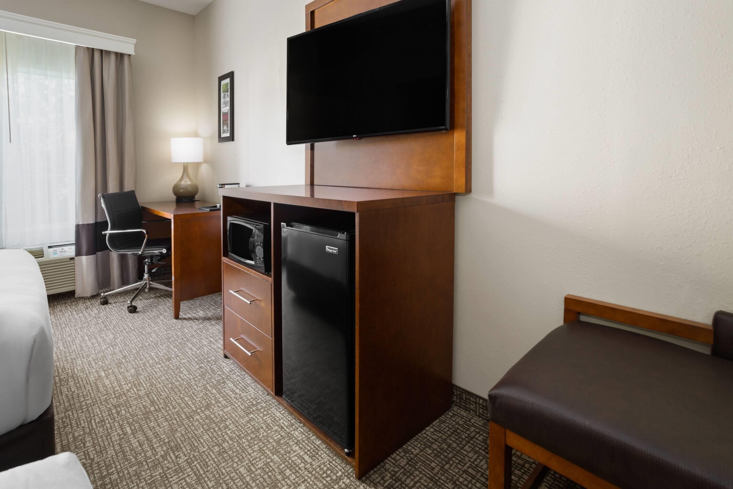A hotel room with a bed, a desk with a chair, a television mounted on the wall, a refrigerator, and a bedside table with a lamp.