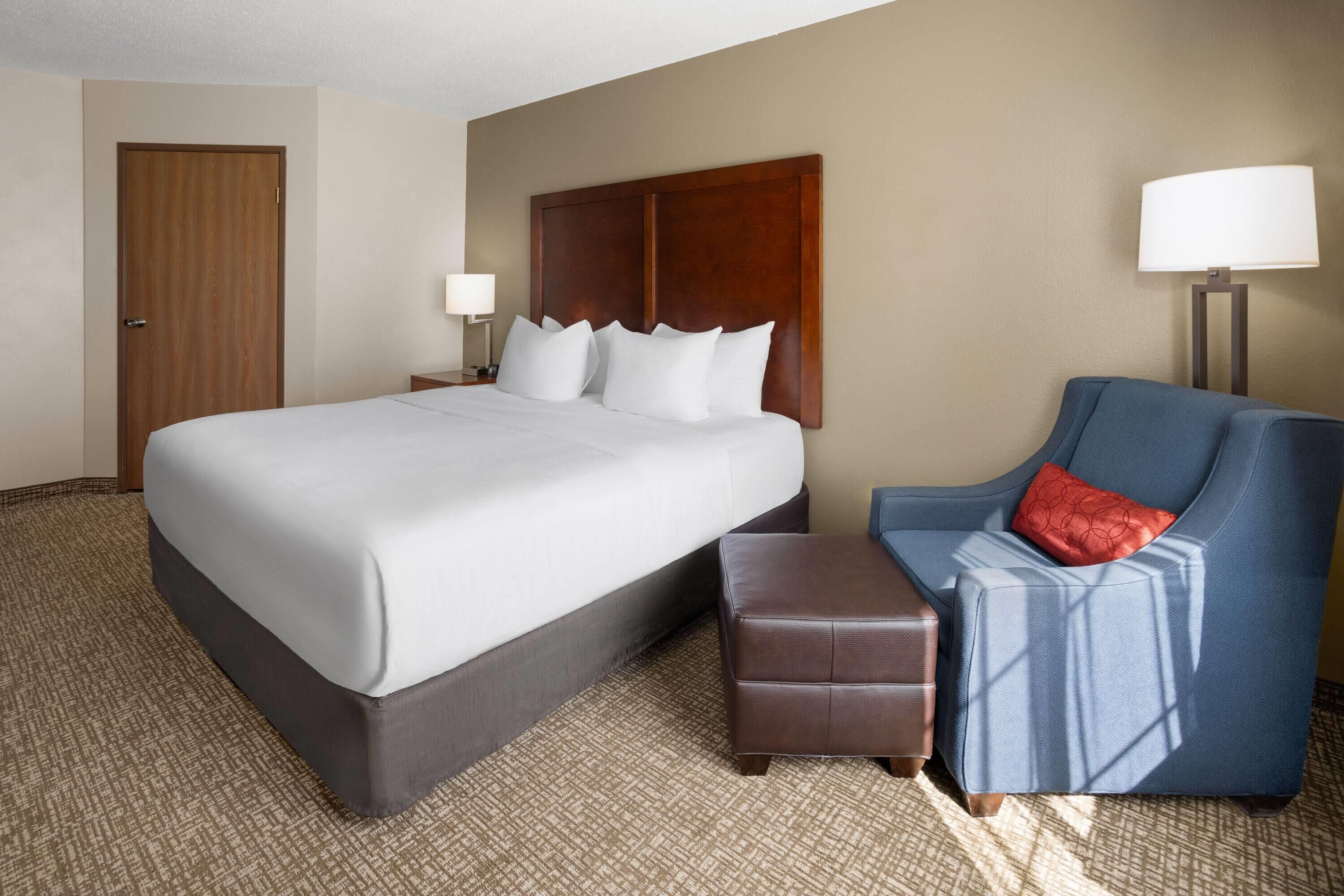 Ein Hotelzimmer mit einem großen Bett, einem blauen Sessel, einem Holzkopfstück, einem Nachttisch mit Lampe und einem gemusterten Teppich.