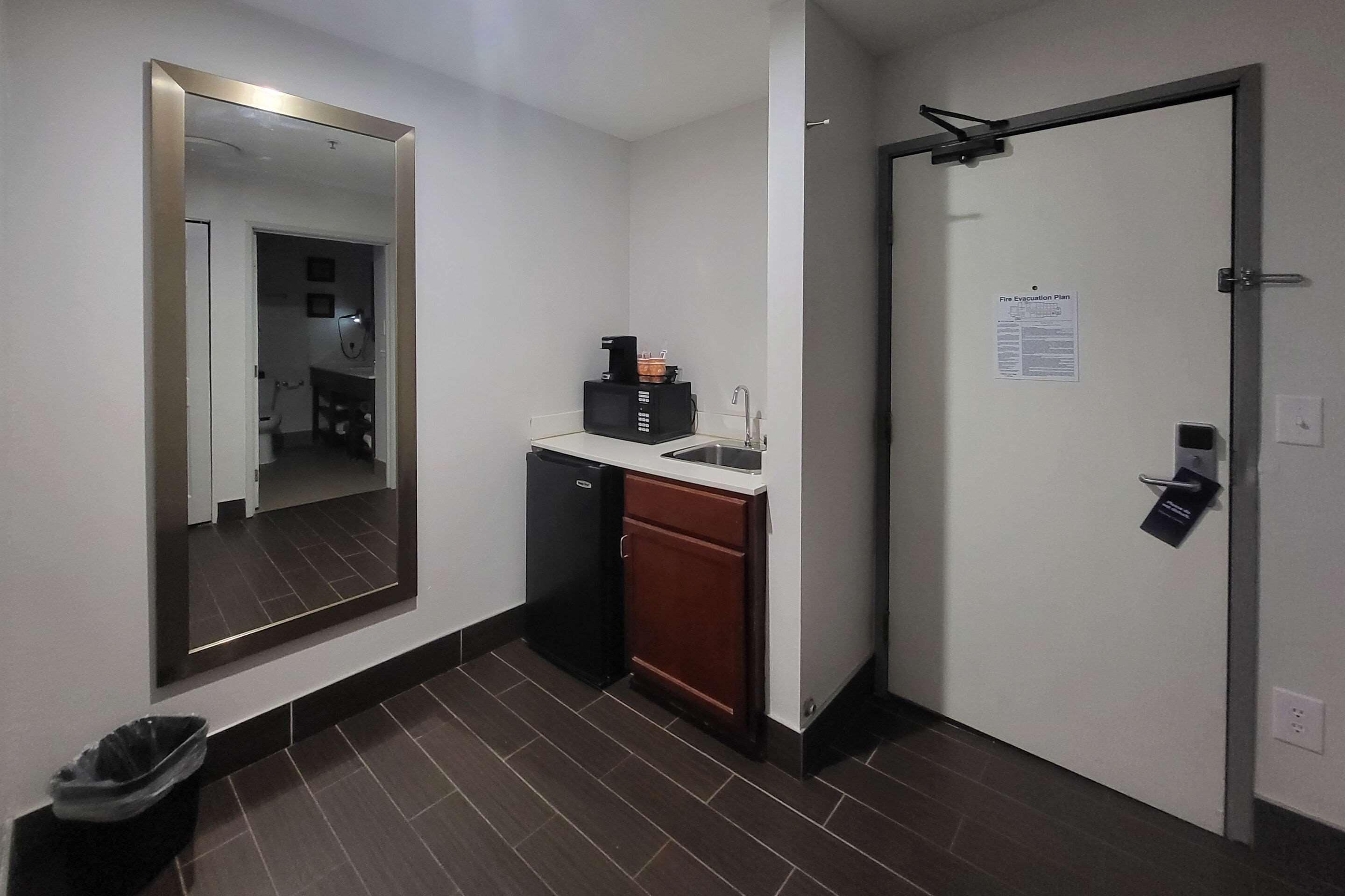 A compact hotel room with a small kitchenette, a mirror, a door with a fire evacuation plan, and a trash bin.