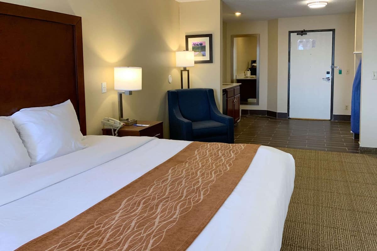 A hotel room with a large bed, a desk with a phone, a chair, a bathroom visible through an open door, and a carpeted floor.