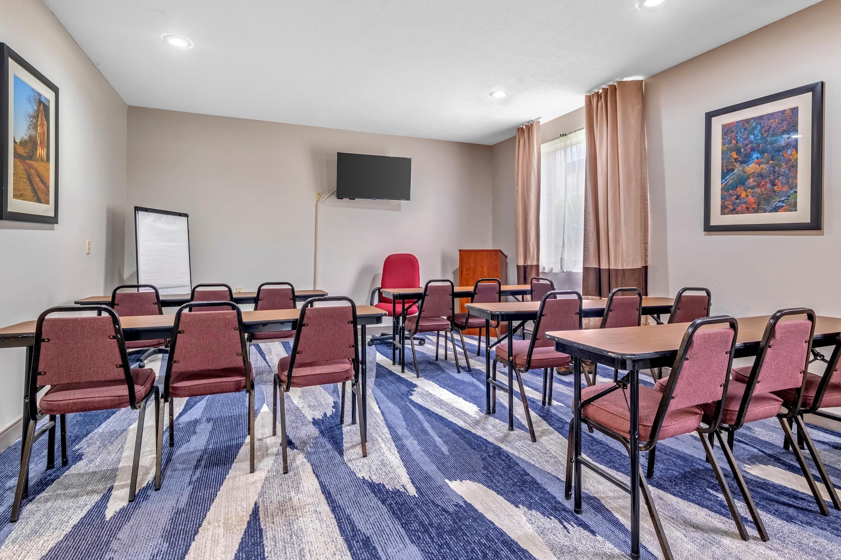 Un salón de conferencias con una alfombra grande, varias mesas y sillas, una pizarra, un televisor y cuadros en las paredes.