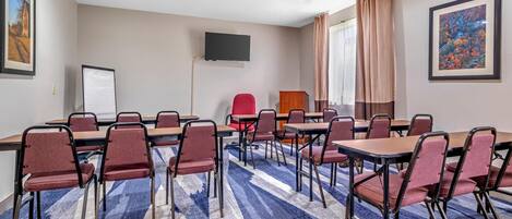 Un salón de conferencias con una alfombra grande, varias mesas y sillas, una pizarra, un televisor y cuadros en las paredes.