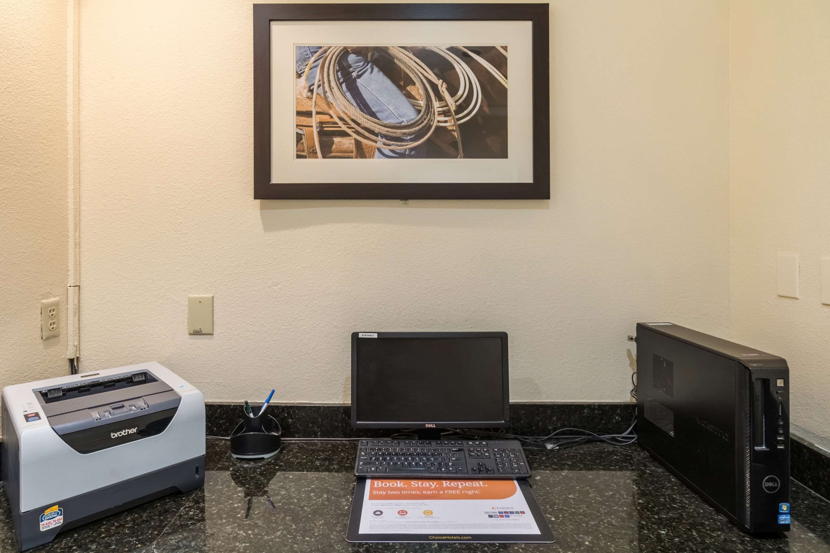 Un escritorio con una impresora Brother, una laptop Dell y una computadora de escritorio Dell. Encima de la pared, cuelga un cuadro enmarcado.