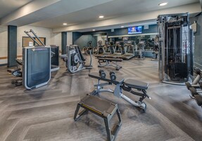 Un gimnasio con cintas de correr, máquinas de pesas y una pantalla de televisión al fondo.