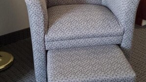 Un fauteuil gris recouvert d’un tissu à motifs, une petite table d’appoint en bois avec un boîte de mouchoirs, et un meuble en bois avec un évier.