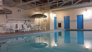 An indoor swimming pool with a tiled floor, a lounge area with white chairs, and a ceiling with exposed beams. - Hastings Country Inn (Hastings)